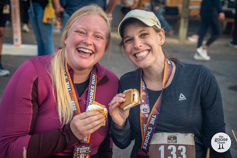 Two runners in The S'mores Run lanyards and medals hold up their s'mores after the race